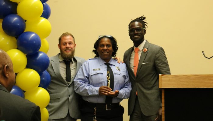 Officer Teresa Palmer, center, standing between William Reed, Community School Coordinator for Cramp Elementary School, and Maxwell Akuamoah-Boateng, Director of Operations for Community Schools, as she holds her Community School champion award trophy. All are smiling.
