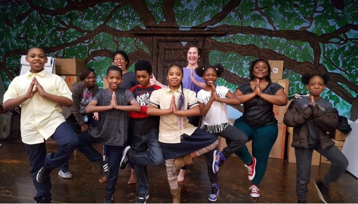 Students and adults stand together in the yoga tree pose. Behind them is a tree mural.