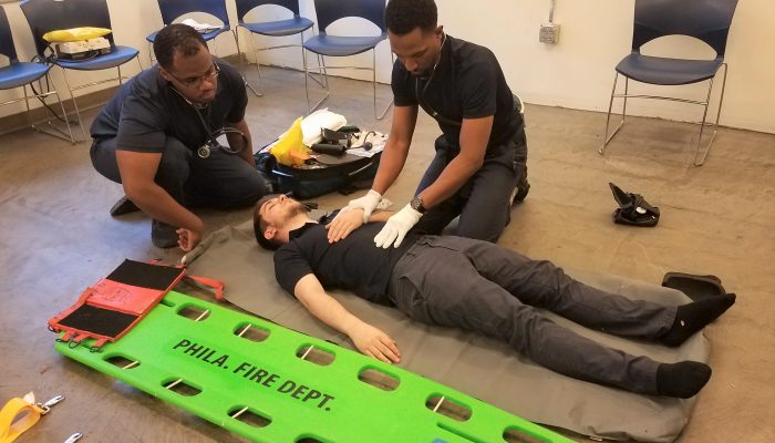 man lying on classroom floor with two men kneeling next to him