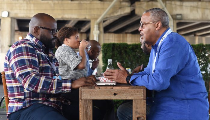 Several people sitting at a table with two men talking to each other.