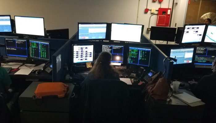 three people sitting at computer consoles with multiple screens