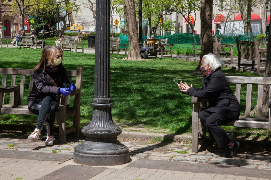 Two people wearing masks talk while sitting on separate benches in a park