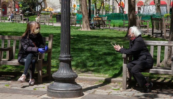 Two people wearing masks talk while sitting on separate benches in a park
