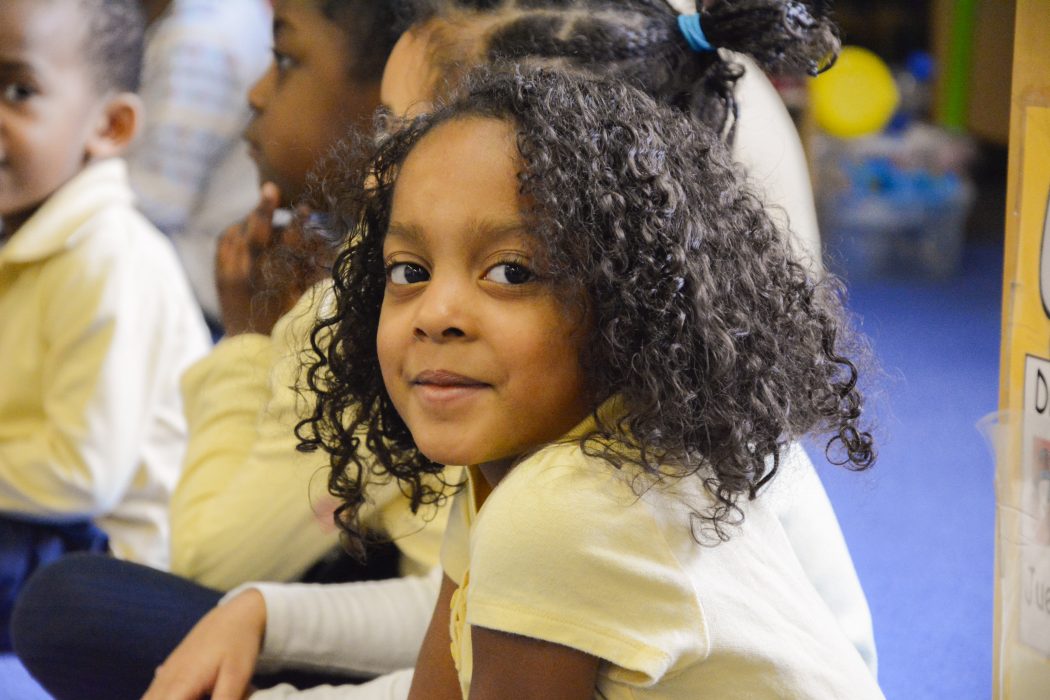 Children gather on the floor for learning. One student is smiling while others pay attention.