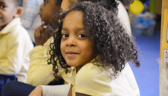 Children gather on the floor for learning. One student is smiling while others pay attention.