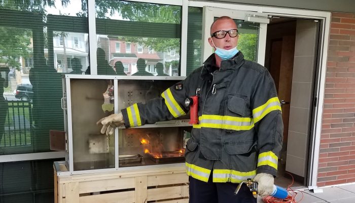 firefighter holding blowtorch and pointing at glass box with flames inside