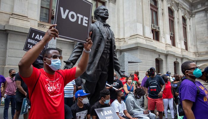 In a crowd near the Octavius Catto statue, a protester wearing a face mask holds up a sign that says "Vote"