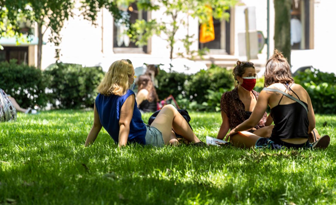 people wearing masks sitting in a small group on the grass in a park