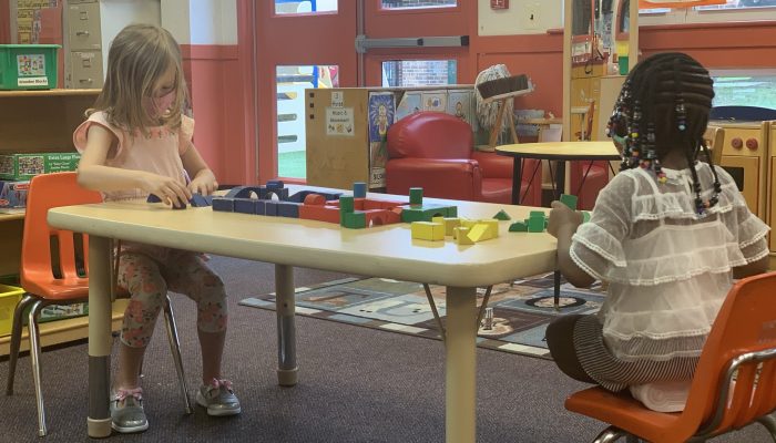 Two PHLpreK students sit together at opposite ends of a table for social distancing. Together they are playing with blocks.