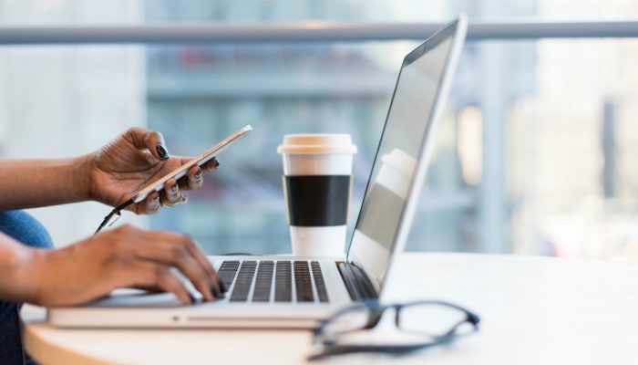 A woman cradles a cell phone in the palm of her hand while she works on a laptop computer