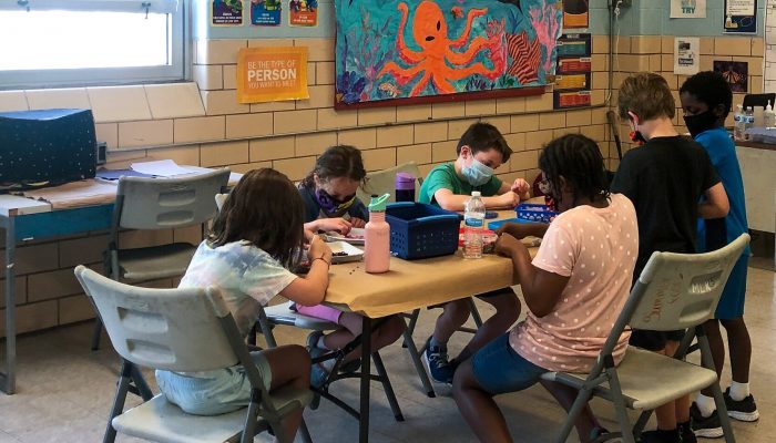 Four students sit together creating crafts at a table inside a Philadelphia recreation center. They are wearing masks and socially distant.
