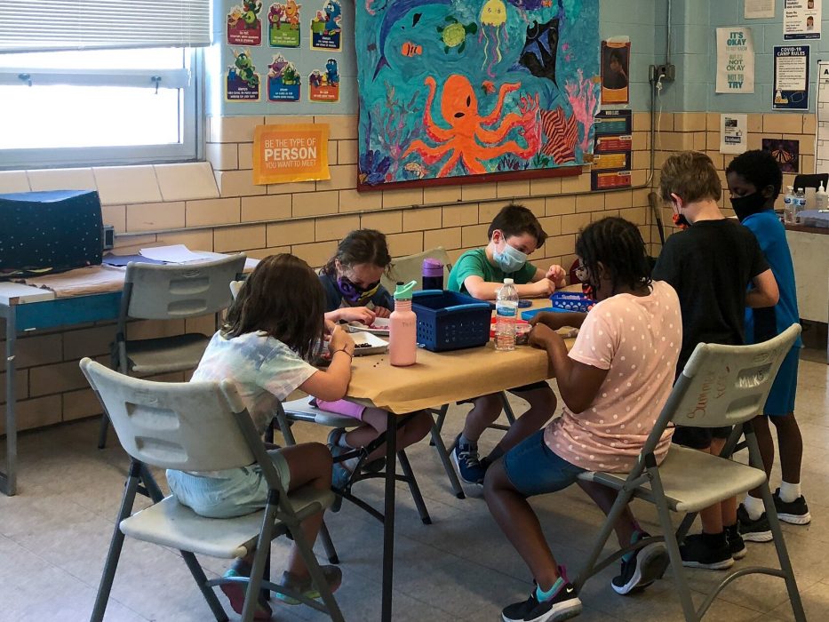 Four students sit together creating crafts at a table inside a Philadelphia recreation center. They are wearing masks and socially distant.
