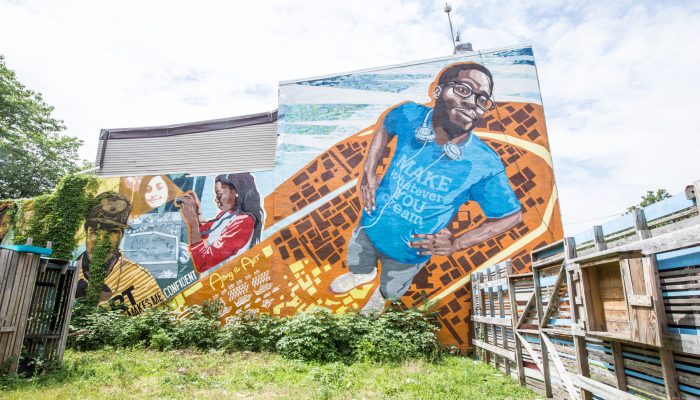 "Art Makes Me Confident" mural, painted on the side of a building. Features several people. The person in the foreground is a young man wearing glasses.