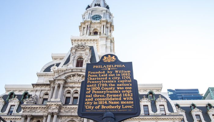City Hall’s clock tower rising in the background behind a historical marker outlining the founding of Philadelphia by William Penn.