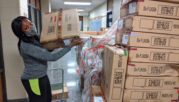 Shira Smillie looks at the camera while holding a brown box full of winter clothes. She has a mask covering the lower half of her face, but looks happy.