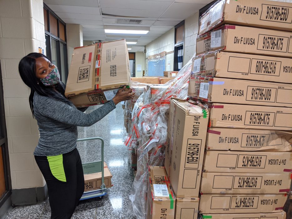 Shira Smillie looks at the camera while holding a brown box full of winter clothes. She has a mask covering the lower half of her face, but looks happy.