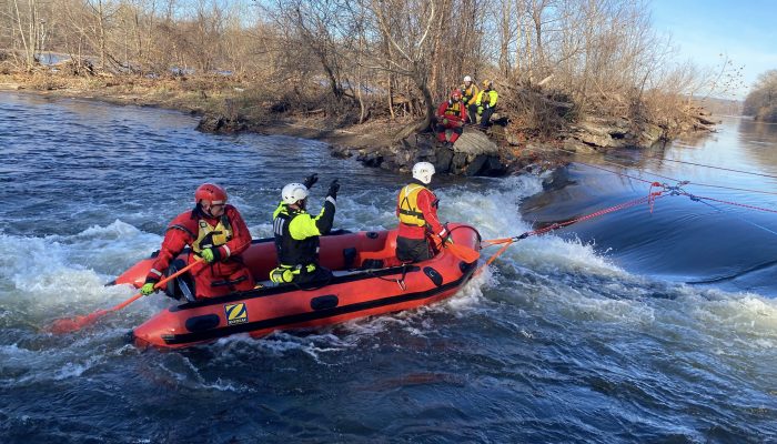 three people in helmets and cold weather gear in inflatable boat tethered to ropes and three people in helmets on rocks on shore