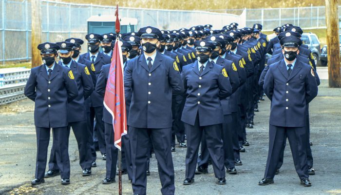 firefighters in dress uniforms and face masks in formation with one holding red flag
