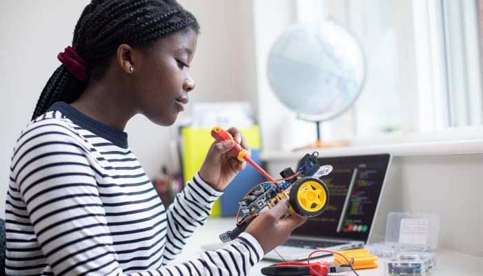 A young girl connects wires on a small machine she is working on as part of her STEM learning program. She is smiling.