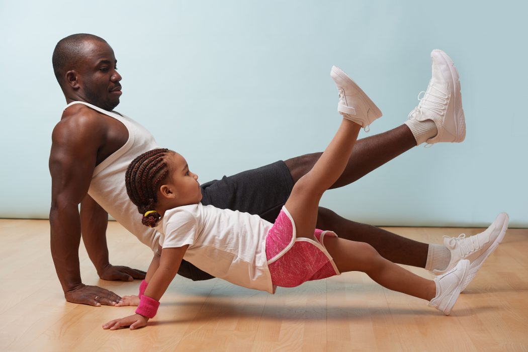 A father and his young daughter exercise on a wooden floor. They are both balancing with their hands on the floor and one foot in the air.