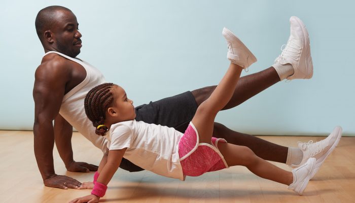 A father and his young daughter exercise on a wooden floor. They are both balancing with their hands on the floor and one foot in the air.