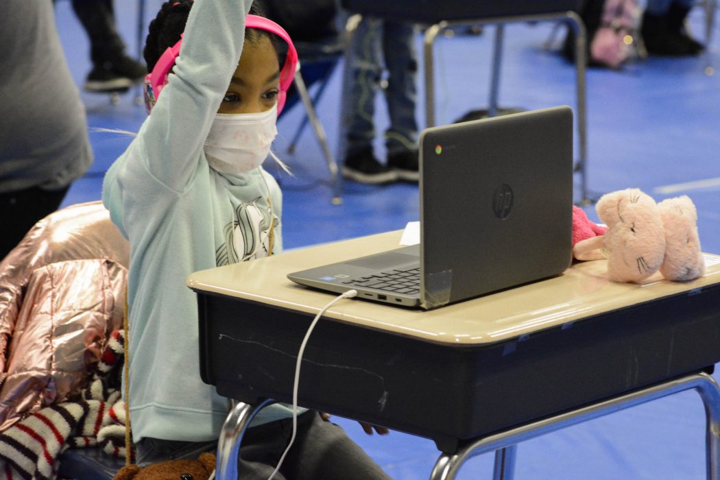 A young girl is seated at a school desk and looking at her laptop. She is wearing headphones and her hand is raised. In the background, additional students appear to be engaged in online learning at individual desks that are spaced out from each other.