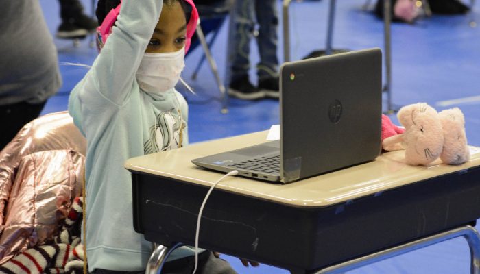 A young girl is seated at a school desk and looking at her laptop. She is wearing headphones and her hand is raised. In the background, additional students appear to be engaged in online learning at individual desks that are spaced out from each other.