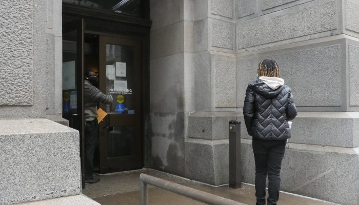 Person waiting outside the City Hall entrance. A man opens the door and motions to her that she can come inside. 