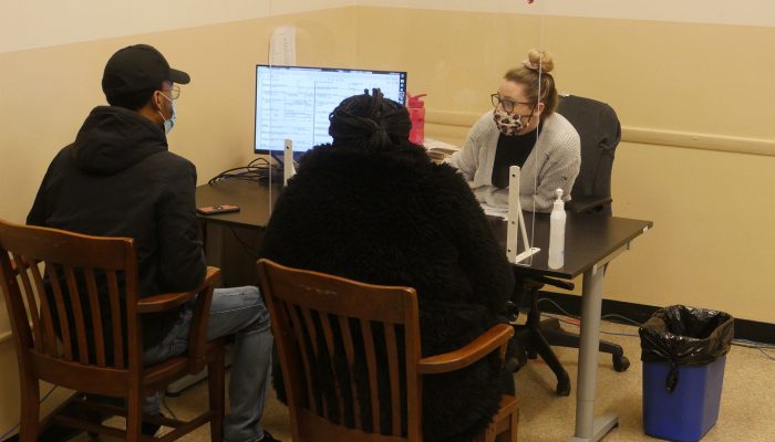 Two residents sitting at a desk in the Marriage License Bureau. A City worker is on a computer on the other side of the desk.