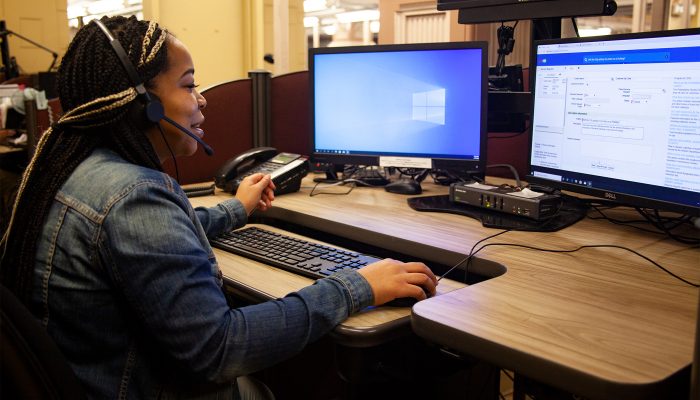 A smiling woman talking into a headset and looking at a computer.