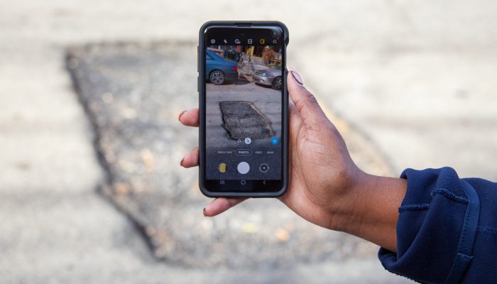 Hand holding a smartphone taking a photo of a pothole on the ground.