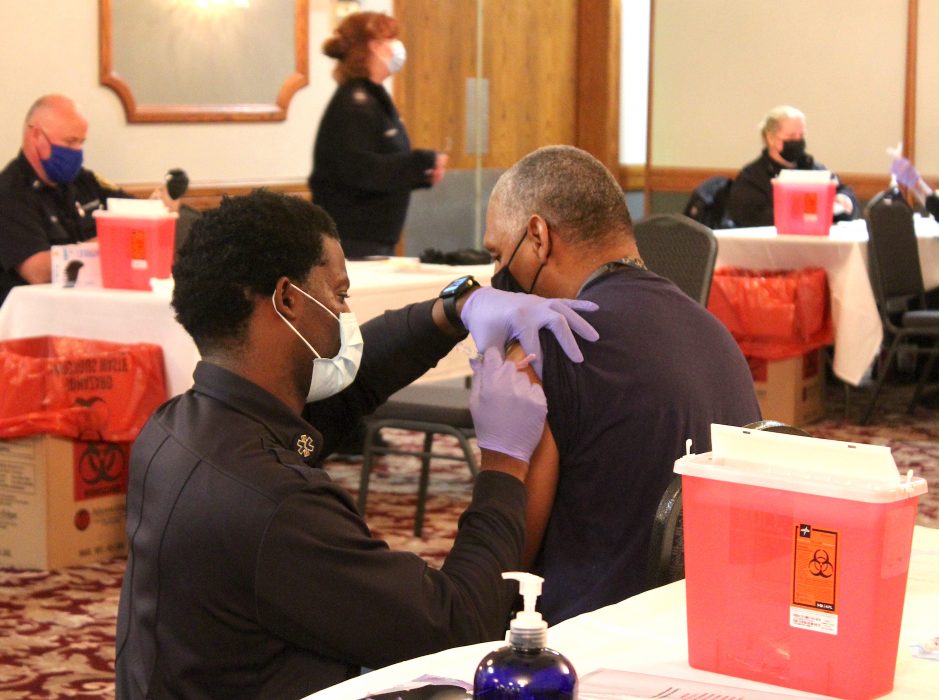 kneeling man in mask and rubber gloves gives shot to seated man in carpeted room