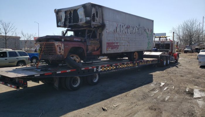 An old, abandoned truck on a long, low-boy tow truck. The old truck is being towed away.