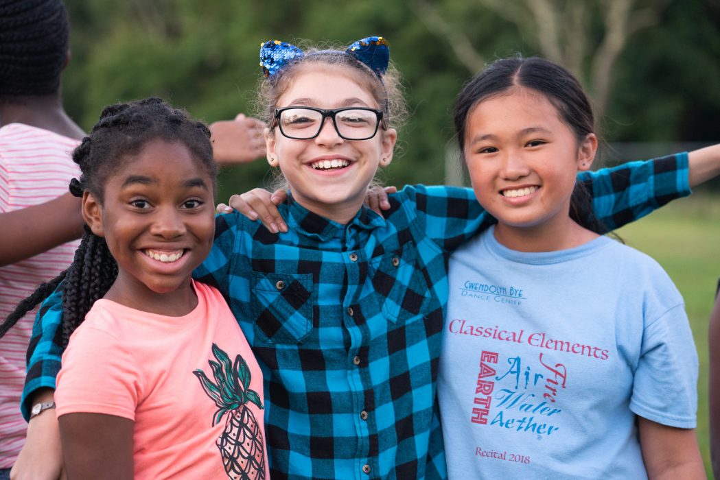 Three school-age girls smiling outside with their arms around each other.