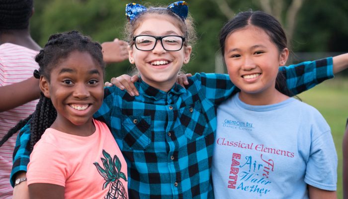 Three school-age girls smiling outside with their arms around each other.