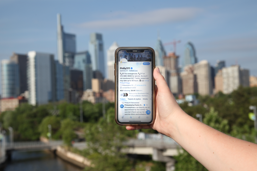 a hand holding a smartphone that has the Philly311 twitter page open. the Philadelphia skyline is in the background