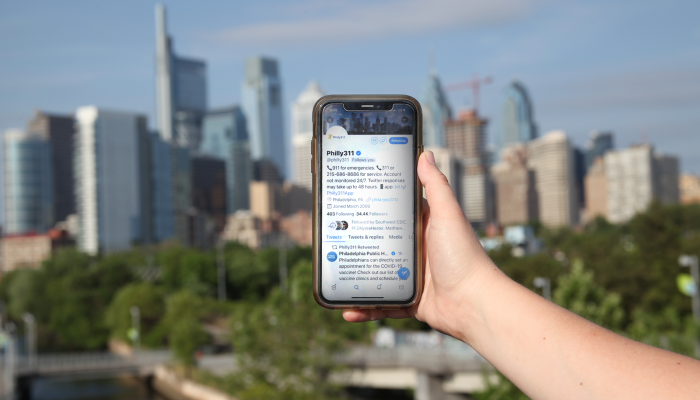 a hand holding a smartphone that has the Philly311 twitter page open. the Philadelphia skyline is in the background