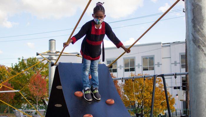 A girl wearing a face mask is standing at the top of a playground structure at a Rebuild site