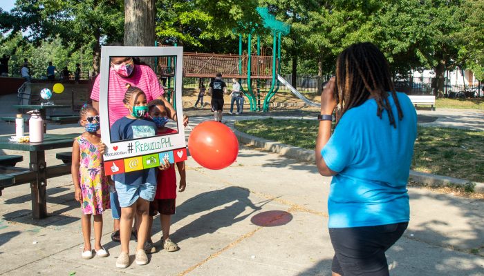 A caregiver and three children pose for a photo at a playground, holding a "#RebuildPHL" prop