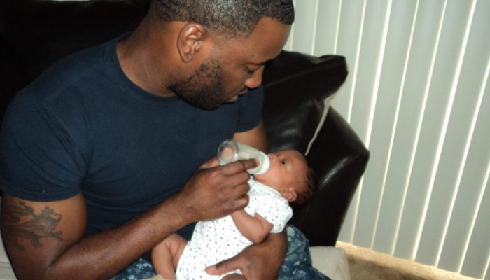 Dad, Branden Kelly, holds his child in his arms. He is feeding the child with a bottle.