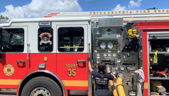 female firefighters stands next to hose attached to side of fire engine in daylight