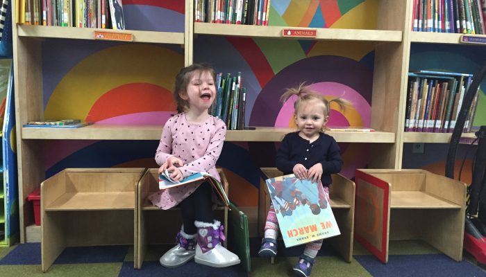 Sara and Abby sit at the library at Western Learning Center. They are smiling and holding books.
