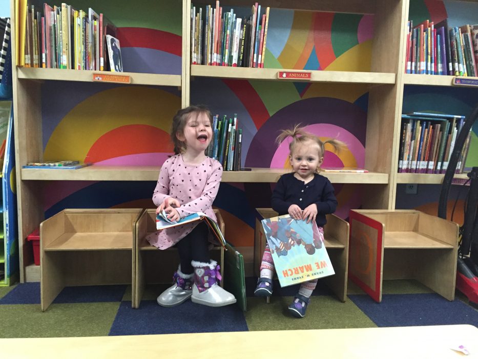 Sara and Abby sit at the library at Western Learning Center. They are smiling and holding books.