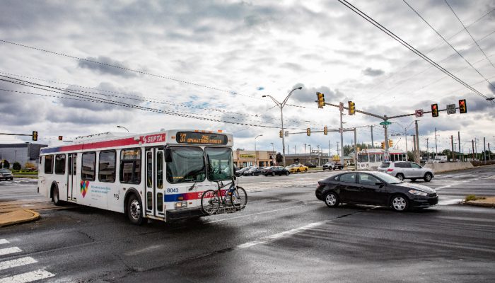 A SEPTA bus is driving on a Philly road on a cloudy day. A black car is to the right and cars are in the background. Traffic lights can also been seen past the black car.