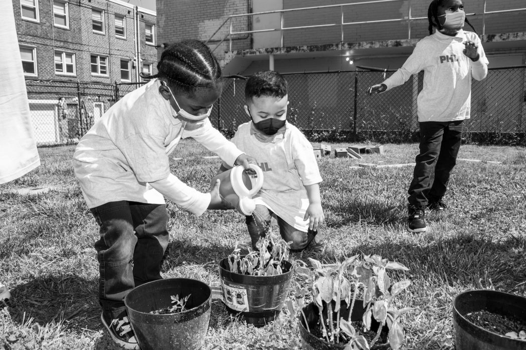 Two phre-K students stand together in front of a plant, they are watering it, working together. Their shirts say PHLpreK and they are wearing face masks.
