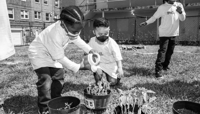 Two phre-K students stand together in front of a plant, they are watering it, working together. Their shirts say PHLpreK and they are wearing face masks.