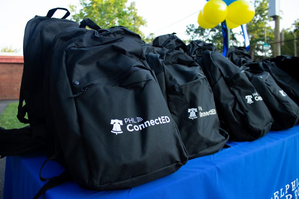 A number of black backpacks with the PHLConnectED logo on them are on a table with a blue table cloth.