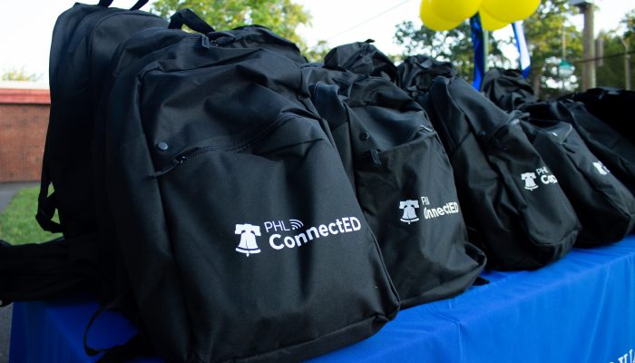 A number of black backpacks with the PHLConnectED logo on them are on a table with a blue table cloth.