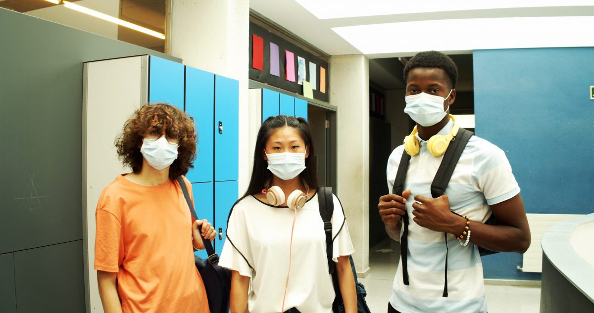 Three students stand together in a hallway with backpacks over their shoulders, they are wearing face masks.