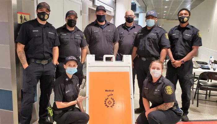 six men in face masks standing and two women in face masks kneeling around sign that says "medical" in english and arabic inside airport terminal